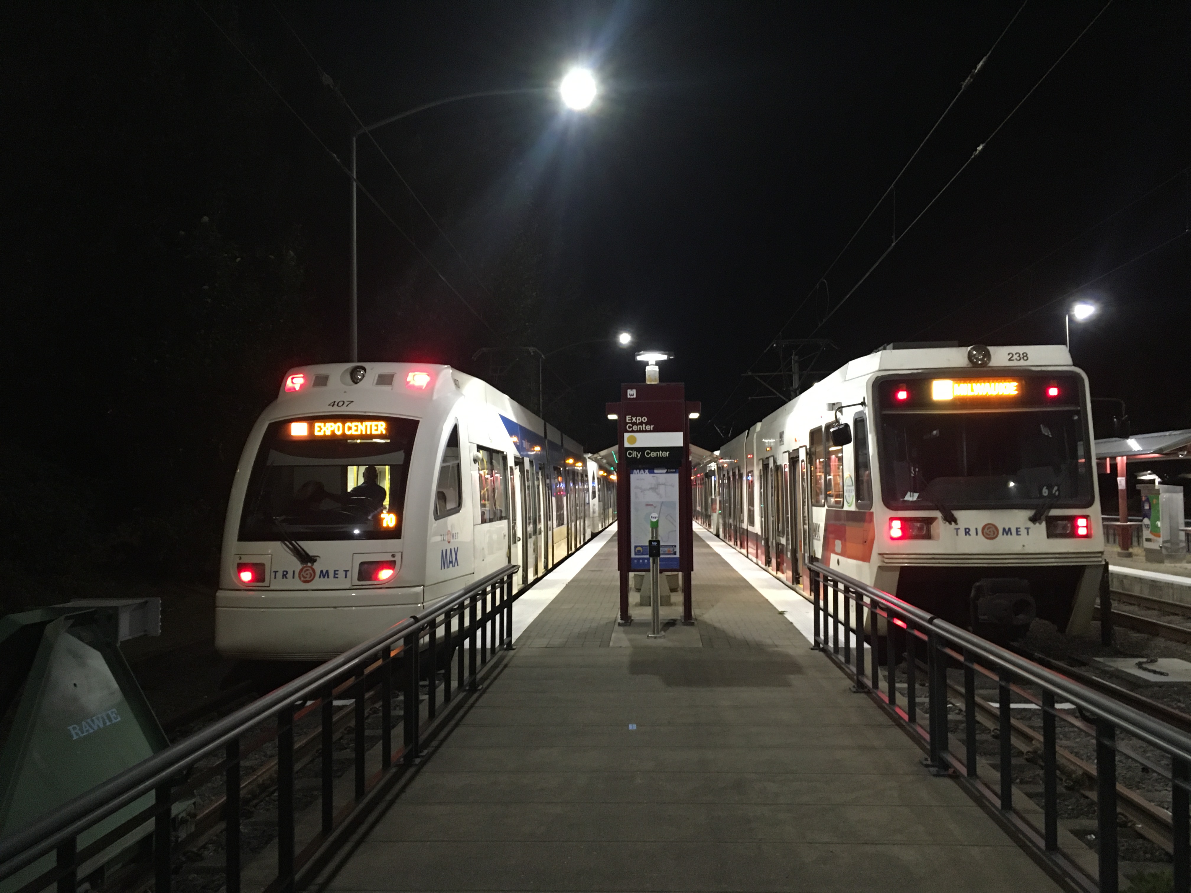 Night-time view of a light rail station, with a newer, sleeker train on the left and and older, boxier train on the right