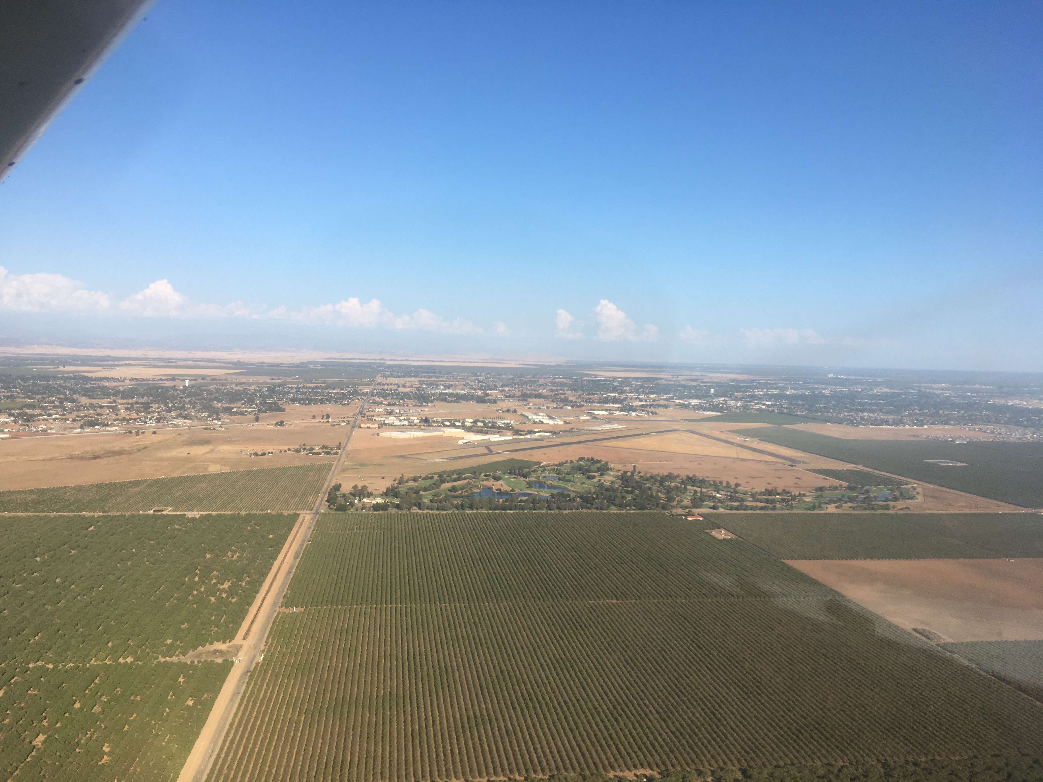 View from the air of wide expans of flat fields, a city with no discernable skyline in the distance, and an airport in the middle ground.  It's Madera, California'